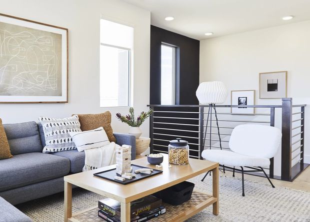 Bright contemporary living room with a grey sectional sofa, layered cushions and throw, light timber coffee table, patterned area rug, sculptural white lounge chair, framed artwork, and a dark stair or mezzanine railing. A compact stair landing and tall narrow windows add architectural interest.