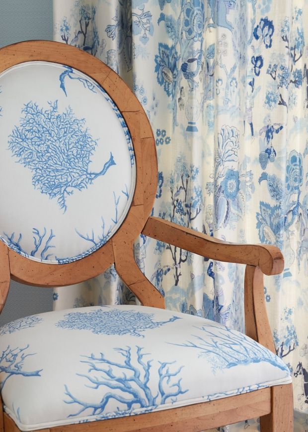 Close-up interior detail of a traditional wooden armchair upholstered in white fabric with blue coral and botanical motifs, positioned against matching blue-and-white floral patterned curtains. The scene has a coordinated coastal-inspired textile scheme and warm timber framing.