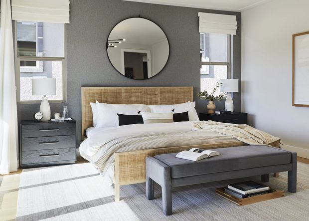 A bright, contemporary bedroom with a woven rattan-style bedhead, layered white bedding, dark bedside cabinets, table lamps, and a large upholstered bench at the foot of the bed. The room features a charcoal textured accent wall, light walls, tall windows with white Roman blinds, and a pale striped rug.