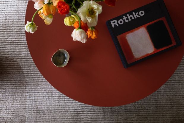 Top-down view of a round terracotta-red side table on a textured grey rug, styled with a small metallic bowl, a loose arrangement of colourful flowers, and a Rothko art book. The scene has a warm, curated interior styling aesthetic.