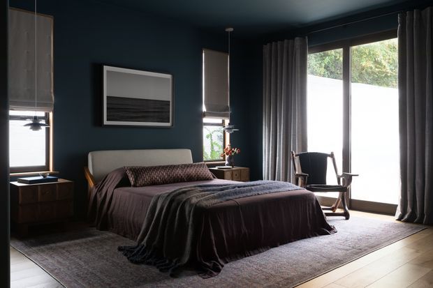 A moody, refined bedroom with deep navy walls and ceiling, a low upholstered bed dressed in layered dark brown and charcoal bedding, dark wood bedside tables, and full-height grey curtains framing expansive glazing. A textured area rug, framed artwork, pendant lighting, and a timber rocking chair complete the calm, sophisticated interior.