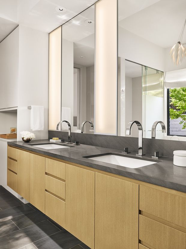 A contemporary double-vanity bathroom with a long floating timber-look vanity, dark grey stone countertop and integrated backsplash, two white rectangular basins, chrome arched mixer taps, and large mirrored wall panels. Vertical illuminated mirror panels add soft ambient lighting. The room has white walls and ceiling, dark large-format floor tiles, recessed downlights, and a sculptural pendant visible at the edge of the frame.
