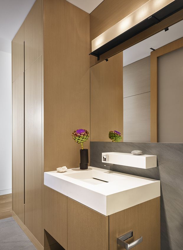 Contemporary bathroom vanity with a floating white integrated basin and wall-mounted white tap fixture, set against grey textured wall panels and extensive warm timber cabinetry. A large mirrored cabinet reflects the room, while concealed linear lighting and black hardware add a refined modern detail. The space is compact, highly tailored, and softly lit.