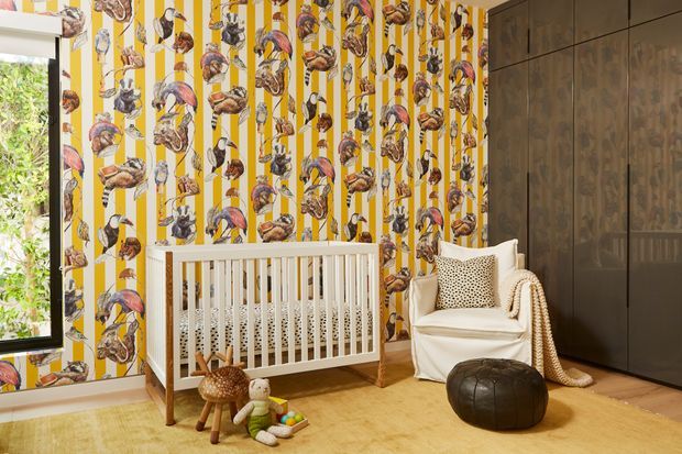 A bright, playful nursery with a white timber cot positioned against bold yellow-and-white animal-patterned wallpaper. The room includes dark full-height built-in cabinetry, a white upholstered armchair with a textured throw, a dark leather ottoman, and a warm light-wood floor. Large glazing at left brings in natural light and views of greenery.