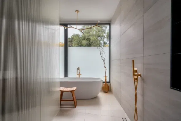 A refined contemporary bathroom with a freestanding white oval bathtub positioned beneath a large window. Pale stone-look wall and floor surfaces create a calm, minimal palette, while a fluted translucent glass partition screens the left side. Brushed brass/gold wall-mounted bath and shower fittings, a sculptural branch in a vase, and a small timber stool add warmth. A black-framed window provides greenery beyond, and a dark wall-mounted screen or mirror is visible at right.