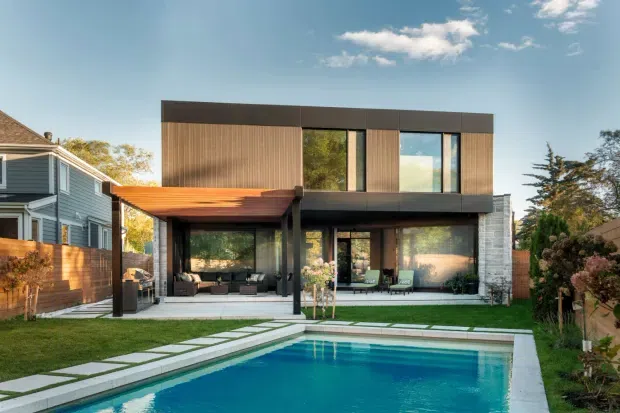 Rear exterior view of a contemporary two-storey residence with a flat roof, dark metal-framed glazing, and warm vertical timber cladding. The house opens onto a covered outdoor entertaining area and a landscaped backyard with a rectangular swimming pool and paved stepping-stone path. Neighbouring suburban homes are visible on either side.