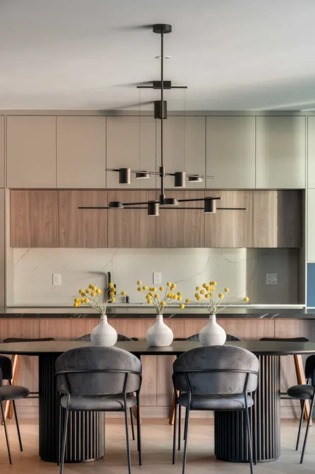 A refined contemporary kitchen and dining area with full-height matte beige cabinetry, warm wood-grain cabinetry, a pale veined stone backsplash, and dark stone benchtops. A long dining table with ribbed cylindrical bases is paired with upholstered charcoal dining chairs, while sculptural black linear pendant lights hang overhead. Three white ceramic vases with yellow branches form a central table arrangement.