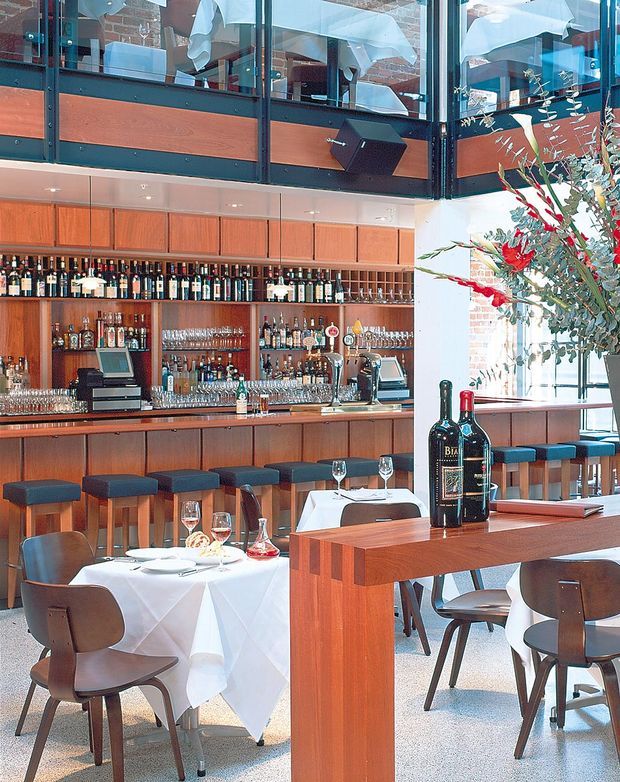 A bright, double-level restaurant interior centered on a timber-clad bar with extensive bottle and glassware display, integrated bar equipment, and a row of upholstered stools. The foreground contains neatly set dining tables with white tablecloths, wine bottles, glasses, and dark timber chairs. A mezzanine level with glass balustrades, exposed brick walls, and industrial-style black framing overlooks the bar. Large glazing and a tall floral arrangement add light and visual interest.