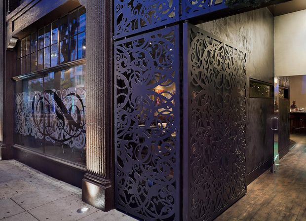 Night-time view of a sophisticated commercial entrance, featuring ornate dark metal laser-cut screens forming a vestibule or partition. Large glazed storefront windows, textured dark facade detailing, warm interior lighting, polished timber-toned flooring, and a bar or hospitality space visible inside create a dramatic, luxurious atmosphere.