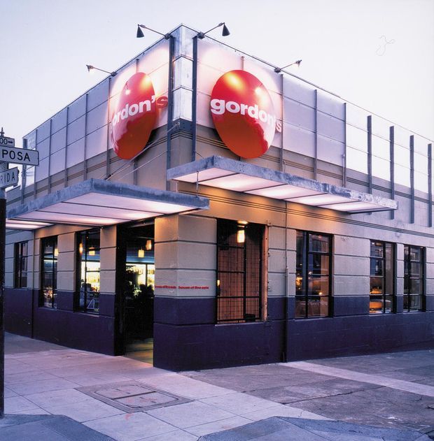 Street-level exterior view of a commercial restaurant building on a corner site. The low-rise facade has pale rectangular wall panels, dark painted lower walls, large windows, projecting metal canopies, exterior wall-mounted lighting, and prominent red circular Gordon's signage. A paved urban footpath and street signs are visible in the foreground.