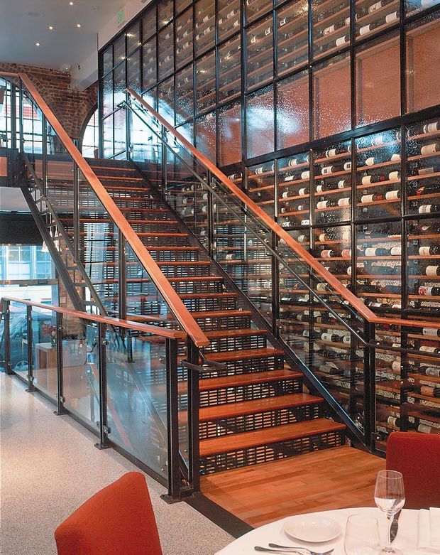 Interior view of a sophisticated multi-level wine-focused commercial or hospitality space. A broad open-riser staircase with dark metal framing, glass balustrades and warm timber handrails rises alongside a floor-to-ceiling glass-fronted wine display wall filled with bottles. Exposed red brick, black steel framing, timber stair treads and warm lighting create an industrial yet refined atmosphere. A dining setting and red upholstered seating are visible in the foreground.