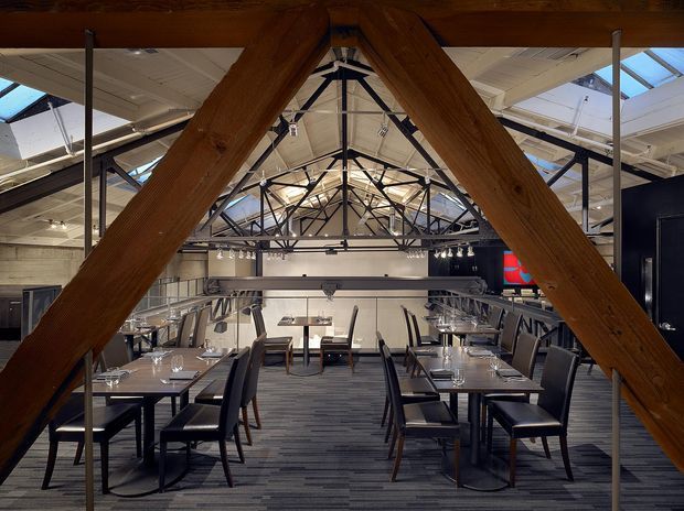 Interior view of a contemporary commercial dining space, likely a restaurant, arranged with multiple neatly set tables and dark upholstered chairs. The room occupies a converted industrial-style structure with prominent exposed timber beams in the foreground, black steel trusses and columns, clerestory-like glazing and skylights, and a mezzanine or raised dining level. The palette is predominantly dark grey, black, white, and warm timber, with strong linear structure and overhead track lighting.