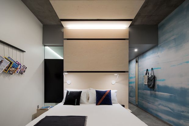 Compact contemporary bedroom with a built-in bedhead and overhead storage, white bedding, dark charcoal cabinetry and ceiling beams, and a distinctive blue-toned wall finish resembling horizontal brushstrokes. A wall-mounted magazine rack and hanging bags add practical details. The room uses integrated lighting and a restrained black, white, grey, and blue palette.