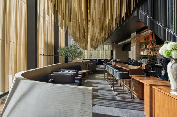 A sophisticated, elongated hospitality interior combining a lounge and bar area. Floor-to-ceiling glazing with sheer curtains lines the left side, while curved upholstered banquettes and lounge seating face a long timber bar with dark stools. A dramatic suspended ceiling installation of numerous slender brass-gold rods creates a canopy overhead. The space also includes dark ceiling surfaces, stone or concrete structural columns, backlit bottle shelving, and warm timber cabinetry, with abundant natural light and a refined contemporary atmosphere.