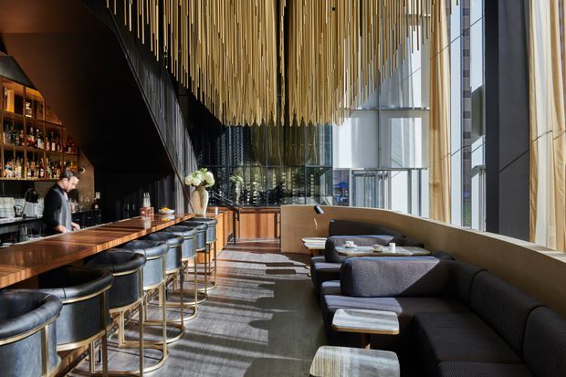 A sophisticated contemporary bar and lounge interior with a long polished timber bar, dark upholstered stools, extensive bottle shelving, curved banquette seating, and low lounge tables. Floor-to-ceiling glazing brings in abundant daylight, while a dramatic installation of suspended slender gold-toned rods creates a striking feature ceiling. The space has a refined hospitality atmosphere with warm timber, dark finishes, and metallic accents.