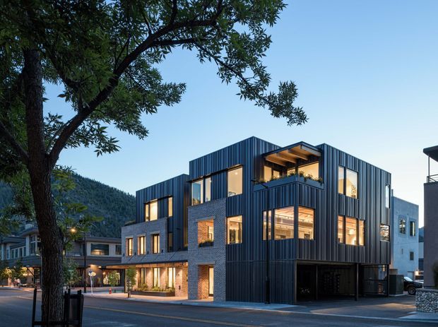 A multi-storey contemporary commercial or mixed-use building on an urban street, set against a wooded mountain slope. The building combines dark vertically ribbed metal cladding, exposed brickwork, extensive glazing, projecting upper-level volumes, and recessed illuminated balconies or terraces. A covered ground-floor opening appears to provide parking or service access, while neighbouring buildings and street activity frame the scene.