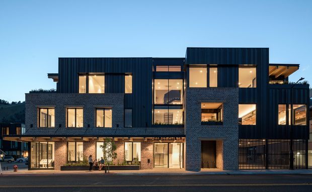 Exterior dusk view of a contemporary multi-storey building with a strongly articulated composition of dark vertical metal cladding, exposed charcoal brick volumes, extensive warm-lit glazing, projecting balconies, and a partially visible timber-lined roof overhang. The building fronts a paved street and includes active ground-level glazed spaces, with a wooded hillside visible in the background.