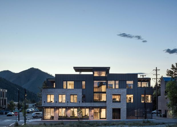 Exterior view of a contemporary multi-storey apartment or townhouse building in an urban mountain setting. The building has a dark metal-clad upper volume, light brick lower facade, extensive rectangular glazing, recessed balconies, and a layered flat-roof composition. Mountains and neighboring buildings are visible in the background.