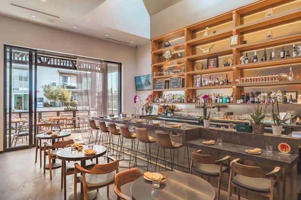 A refined contemporary bar and dining space with a long stone bar counter, timber shelving illuminated with integrated warm lighting, extensive bottle and glassware displays, and a mix of high stools and small round dining tables. Full-height glazed doors open to an outdoor terrace, while a high ceiling and neutral finishes create an airy hospitality atmosphere.