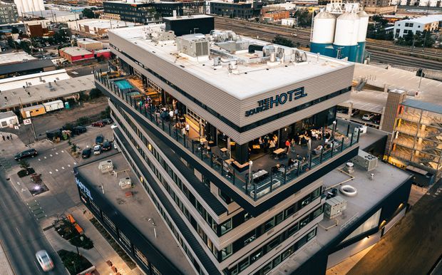 Aerial view of a contemporary multi-storey hotel building in an urban industrial precinct. The building has a flat roof with extensive rooftop mechanical equipment, horizontal metal cladding, large glazed façades, and a prominent elevated outdoor terrace with dining and lounge seating. Surrounding warehouses, industrial tanks, roads, and parking areas are visible.