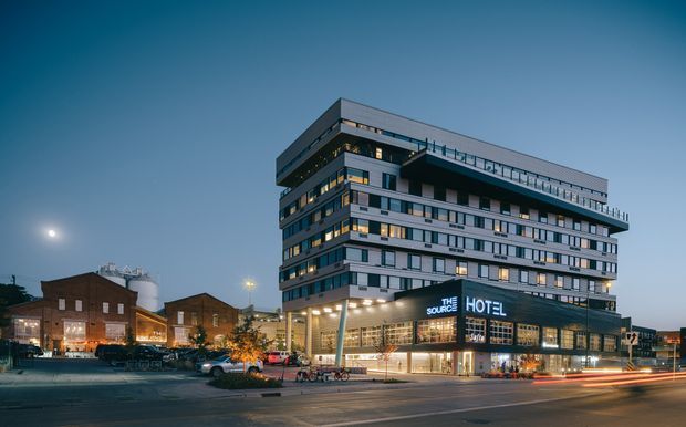 Twilight exterior view of a contemporary multi-storey hotel building with layered projecting floor plates, extensive strip windows, illuminated ground-floor glazing, and a prominent hotel sign. Older brick industrial-style buildings and a broad urban street sit in the surrounding streetscape.