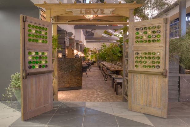 An inviting entrance to a lush, open-air restaurant or hospitality space. Wide timber-framed double doors feature decorative panels inset with rows of green glass elements, opening onto a paved courtyard-like dining area with long communal tables, chairs, planting, and a rustic bar or service counter. Timber beams and a partially glazed or open roof structure create a warm, informal atmosphere.