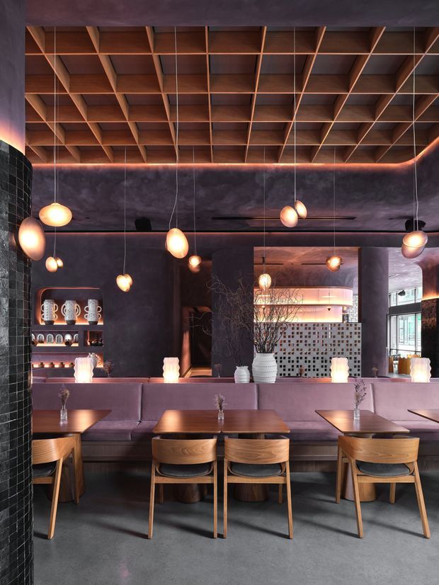 A sophisticated dark-toned restaurant or hospitality dining interior with mauve upholstered banquette seating, timber dining tables and chairs, and a dramatic coffered timber ceiling. Numerous suspended globe pendant lights create warm pools of light against charcoal textured walls, while a large mirror opens the view toward a tiled service or bar area. The space combines rich timber, polished concrete flooring, dark surfaces, and warm ambient lighting in an atmospheric contemporary fit-out.