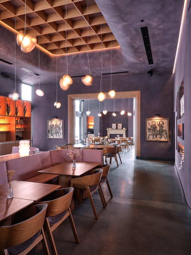 A stylish restaurant dining interior with warm, atmospheric lighting. The space features rows of timber dining tables and curved-back wooden chairs, upholstered banquette seating, a dark polished concrete floor, deep mauve plaster walls, illuminated timber shelving, framed artwork, and numerous suspended globe pendant lights. A distinctive exposed timber coffered ceiling adds strong architectural character, while a large opening leads to a second dining area.