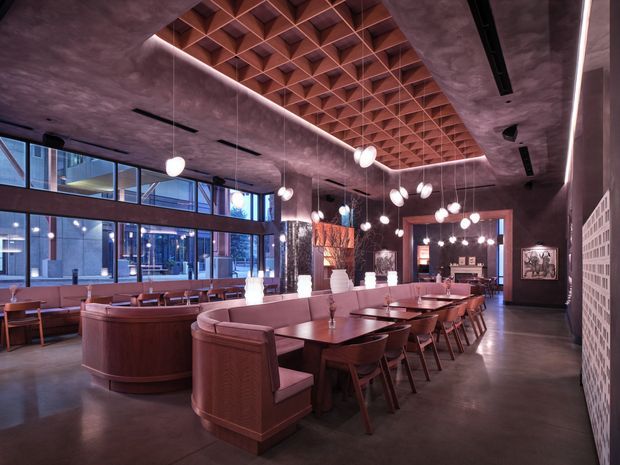 A spacious upscale restaurant or hospitality dining interior with long communal timber dining tables, upholstered banquette seating, curved booth seating, and numerous spherical pendant lights. The room features polished concrete flooring, dark textured walls and ceiling, expansive glazing along one side, and a striking geometric timber coffered ceiling feature in warm reddish-brown tones. A perforated masonry or breeze-block partition is visible on the right, contributing to the contemporary industrial character.