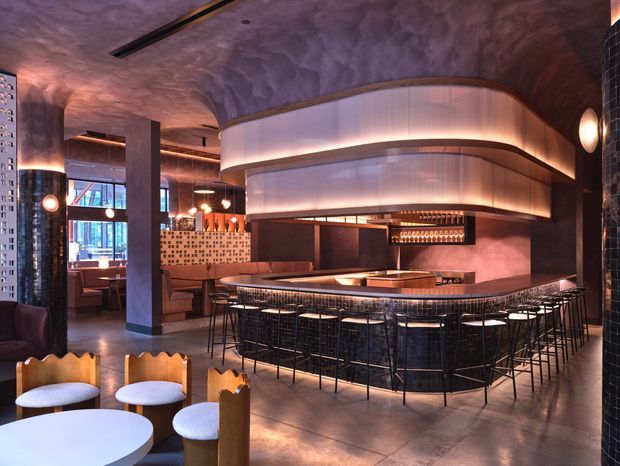 A dramatic, sophisticated hospitality interior featuring a large curved bar with dark stone or tile cladding, extensive high stools, and a glowing translucent curved overhead canopy. The space has a textured sculptural ceiling, dark columns, warm ambient lighting, decorative pendant and wall lights, booth seating, and patterned tile accents. The palette is predominantly dark brown, charcoal, bronze, and warm amber, creating an atmospheric upscale bar or restaurant setting.