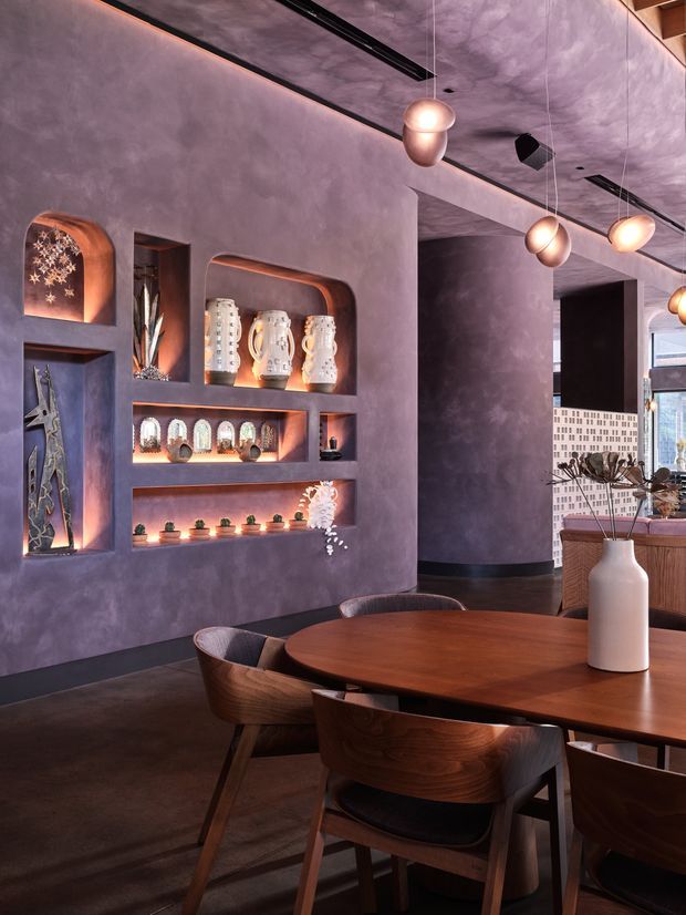 A sophisticated dining area with a large rounded timber dining table and sculptural timber chairs. The room features dramatic mauve-purple textured walls, integrated recessed display niches with warm lighting, decorative ceramics, dark flooring, and multiple glowing pendant lights. A bar or lounge area is partially visible beyond the dining space, contributing to an upscale hospitality-style atmosphere.