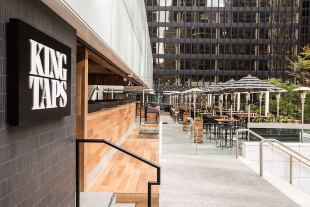 Exterior view of the King Taps commercial hospitality venue opening onto a large urban terrace. The scene features a dark tiled facade with prominent signage, warm timber-clad service frontage, broad concrete paving, outdoor dining tables beneath repeated umbrellas, railings, planters, and a high-rise office building backdrop.