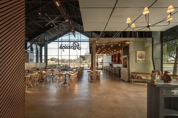 A spacious contemporary restaurant or hospitality interior with an open dining area, high industrial-style exposed roof structure, extensive floor-to-ceiling glazing, and a polished concrete floor. Numerous timber dining tables and chairs fill the space. A bar and service counter sit toward the rear, while decorative pendant and chandelier lighting, framed artwork, and a dark slatted feature wall add visual detail. The view through the glazing shows an exterior setting but the primary subject is the commercial dining interior.