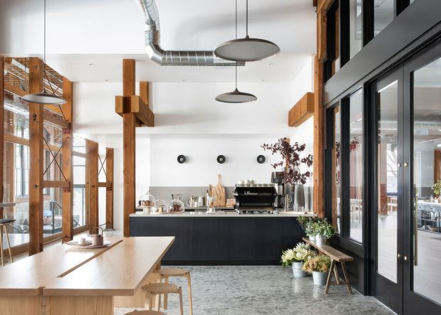 A bright, open-plan kitchen and dining space with a large black island, integrated coffee station, timber structural posts and beams, exposed metal ductwork, polished concrete flooring, and extensive black-framed glazing. A long timber dining table occupies the foreground, while industrial pendant lights and warm timber details create a contemporary warehouse-inspired interior.