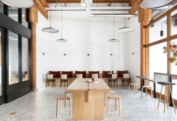 Bright contemporary café or communal dining space with a long light-wood central table, stools, small tables and chairs, and a built-in burgundy banquette along a white wall. The interior features exposed timber beams, exposed services and ductwork, large black-framed glazed doors and windows, polished pale flooring, and suspended oval pendant lights. A dark framed glazed room or entry zone is visible at left.