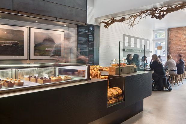 A bright contemporary café or bakery interior with black textured service counters, illuminated glass pastry display cases, wall-mounted artwork, and a mirrored rear counter. Customers sit on high stools beside a white horizontally lined wall, exposed brick, and large windows. A striking organic branch-like sculptural installation spans the ceiling, adding a natural accent to the refined commercial fit-out.