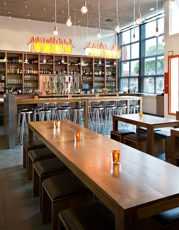 A bright contemporary bar and dining interior with a long timber communal table surrounded by dark upholstered benches and stools. The rear bar features extensive open bottle shelving, a timber counter, beer taps, and colourful sculptural pendant lights. Large industrial-style black-framed windows bring in abundant daylight, while exposed-looking acoustic ceiling tiles and numerous hanging bulb lights contribute to the lively hospitality atmosphere.