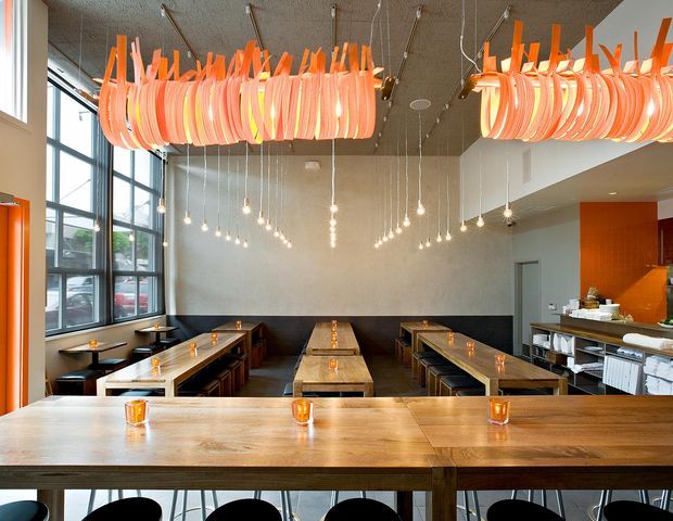 A contemporary restaurant or café interior viewed from behind a large timber communal counter. The space features long timber dining tables with dark banquette seating, extensive floor-to-ceiling glazing along the left wall, a service counter and open kitchen area at right, and dramatic suspended orange sculptural pendant lights combined with rows of small hanging bulbs. The palette is warm timber, charcoal, grey, and vivid orange, creating a lively but refined dining atmosphere.