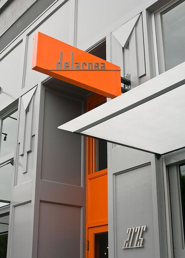 Close-up exterior view of a modern commercial or institutional building entrance. The facade uses light grey panels with angular folded metal detailing, dark-framed glazing, and a prominent vivid orange projecting canopy and orange entry door. Metallic signage is mounted above and beside the entrance.