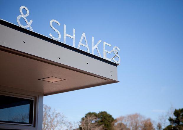 Close-up exterior view of a modern low-profile building canopy with large raised “& SHAKES” signage along the roof edge. The facade and soffit are light-colored with dark trim, and a glazed opening is partially visible below. Clear blue sky and trees form the background.