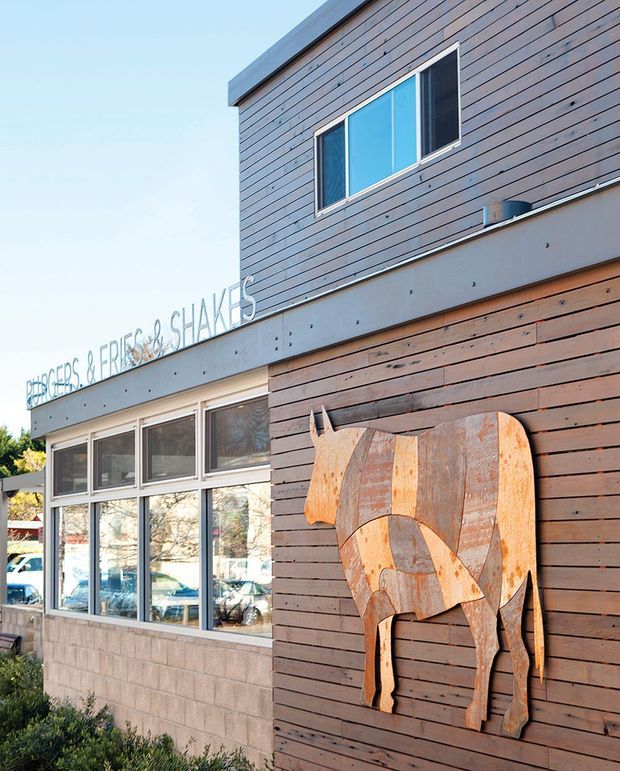 Exterior view of a contemporary two-storey commercial building with horizontal dark timber cladding, a light concrete or rendered band, large ground-floor windows, and a projecting sign reading “Riders, Reapers & Shakes.” A large decorative horse silhouette is mounted on the timber-clad facade. The scene is brightly lit with a clear sky and some street-side planting visible.