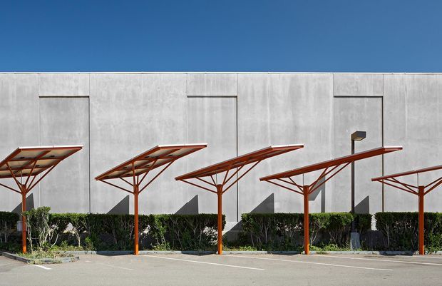Exterior view of a contemporary commercial or industrial building with a long, pale grey concrete facade, narrow recessed vertical openings, and a row of angular rust-red steel shade canopies over a mostly empty paved parking area. Low hedging and landscaping line the base of the building beneath a clear blue sky.