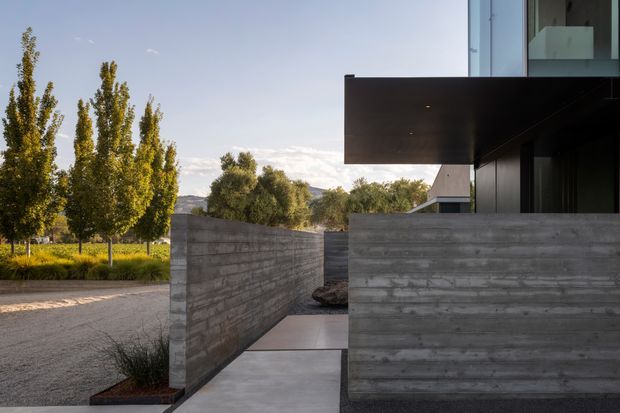 Exterior architectural view of a contemporary residence, showing a concrete-walled entry path, dark cantilevered upper volume, extensive glazing, and a landscaped rural setting with tall trees and distant hills.