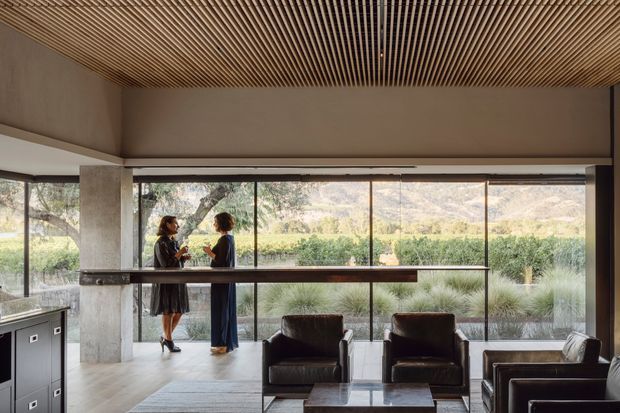 A refined contemporary interior with a large glazed wall opening onto landscaped grounds and distant mountains. Dark leather lounge chairs and a low coffee table sit beneath a warm timber slatted ceiling, while a long dark counter or console runs along the glazing. Exposed concrete columns and neutral finishes create a calm, sophisticated lounge or reception-like setting.