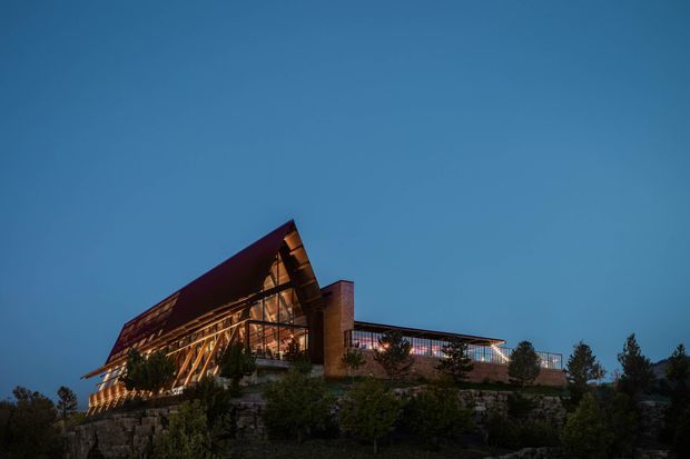 Twilight exterior view of a dramatic contemporary building set on a landscaped, rocky slope. The structure features a steep angular gabled roof, extensive warm-lit glazing, exposed timber-like structural framing, and a long low glazed wing extending to the right. Trees and stone retaining elements surround the building.