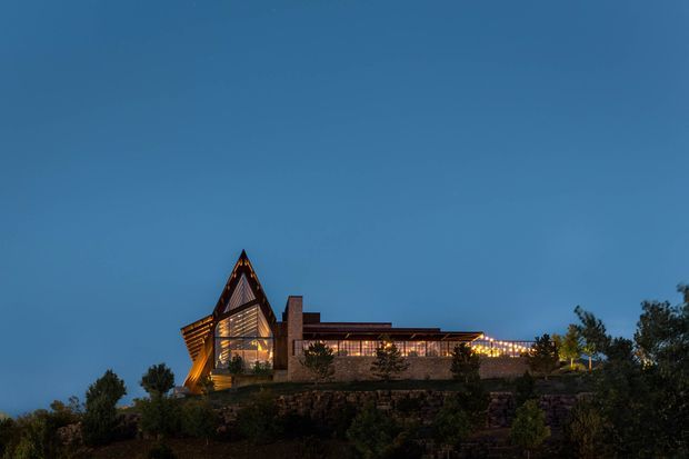 Dusk exterior view of a distinctive large residence or lodge perched on a landscaped, elevated site. The building features a dramatic steep triangular roof form with illuminated timber-like detailing, extensive warm-lit glazing, a long low wing, and a prominent masonry chimney or vertical wall element. Trees and planting frame the foreground against a deep blue evening sky.
