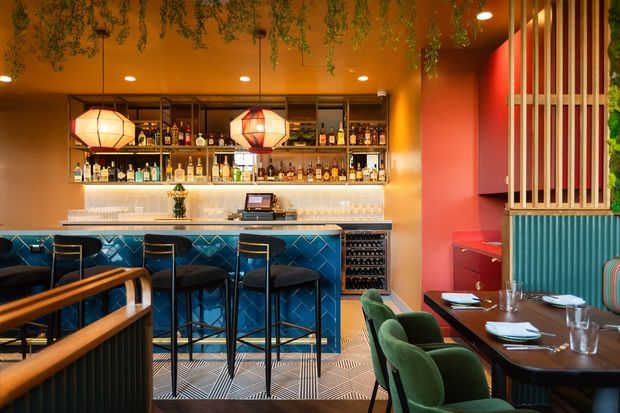 Vibrant, richly coloured commercial bar and dining interior with a long blue-tiled bar, black bar stools, illuminated bottle shelving, geometric pendant lights, and hanging greenery. A dining area with dark tables and green upholstered seating occupies the foreground, while timber vertical slats and ribbed wall panelling add architectural detail.