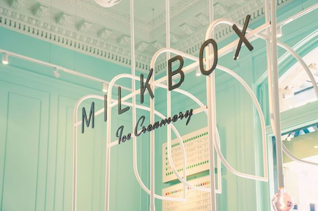 Interior view of a pastel mint-green ice cream shop with ornate traditional ceiling mouldings and wall panelling. A white tubular metal display structure forms looping arches around large black lettering spelling “MILKBOX” and script “Ice Creamery,” with a brightly lit service or display area visible at the right.