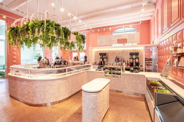 A brightly lit, highly stylised commercial food and beverage interior with a curved service counter, central display island, coffee and drink preparation stations, refrigerated product displays, and menu boards. The space features extensive coral-pink geometric wall panelling, pale textured tile cladding on the counters, terrazzo-style flooring, white exposed services and track lighting, and abundant hanging greenery.