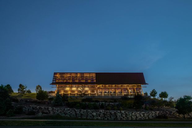Dusk exterior view of a long, elevated modern building set on a landscaped, rocky site. The structure has a dramatic steeply sloping roof with warm illuminated glazing and timber-like structural elements beneath, creating a strong horizontal profile against the blue evening sky.